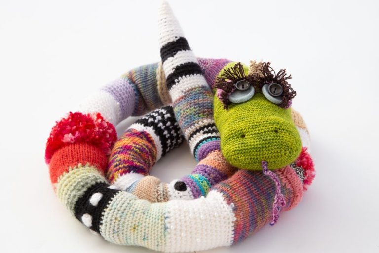 quietschvergnügte gestrickte Schlange, die sich eingerollt hat und nun mit ihren langen Wimpern und großen Knopfaugen dir zuzwinkert