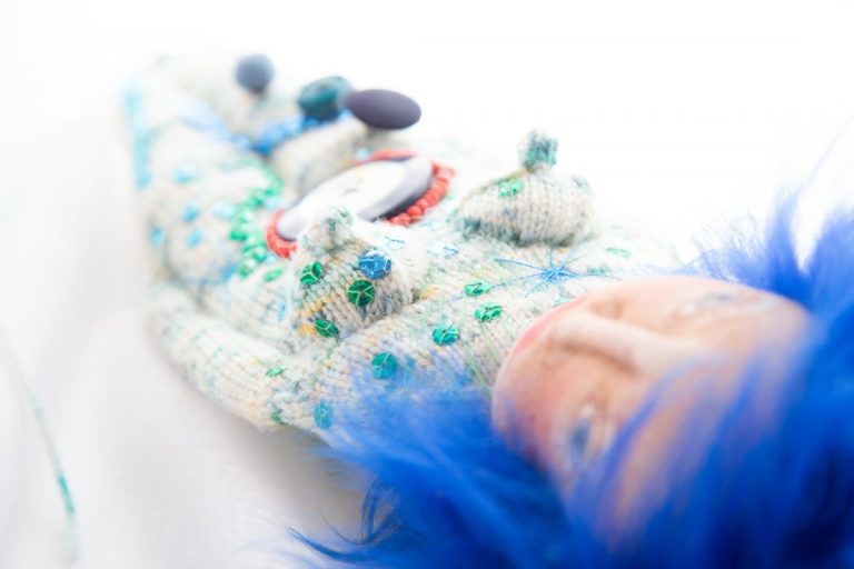 blauhaariges Wesen mit weiß-türkisfarbenem Strickkörper, bestickt mit Pailletten in Grün, Blau und Türkis, stilisierte Yoni, die mit roten Perlen umstickt ist