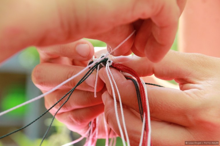 zwei Paar Hände knüpfen eine Schnur aus weißen, grünen und roten Fäden