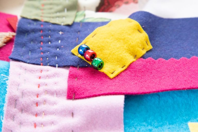 eine blaue, rote und grüne Plastikperle auf gelbem Rechteck, das alles auf Ebenen von vielen anderen Farben, handgenähter Quilt