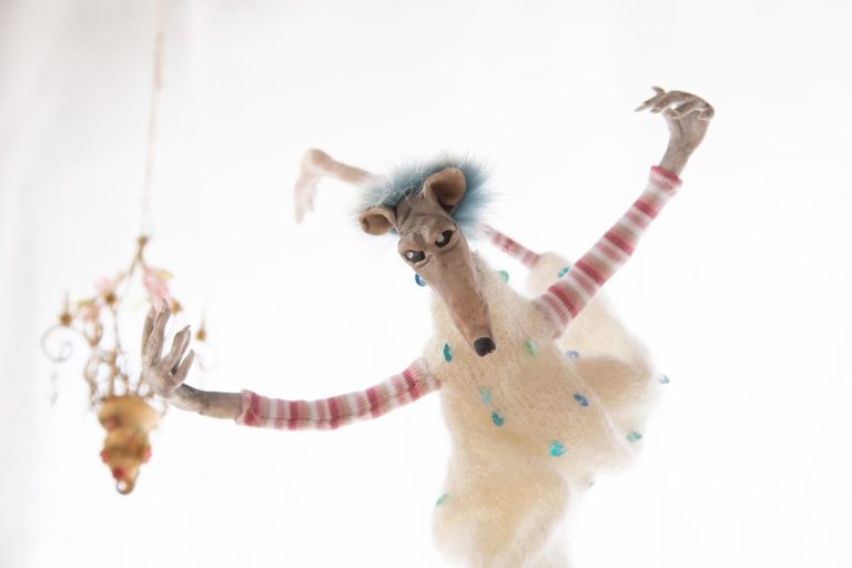 filigrane, tanzende Ratte, Arme sind oben, ein Bein nach hinten hinaus gestreckt, der vordere Fuß auf Zehenspitzen und durchgestreckt, vorherrschende Grundfarben Weiß, Türkis und Rosatöne