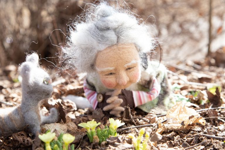 Emely, angegraute Hippie-Frau, gefilzt, liegt vor den diesjährigen gelben Primeln, die aus der Erde schauen, gut bewacht von ihrem Kater