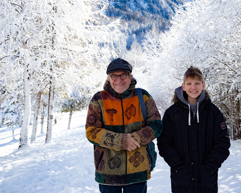 Zwei Männer, ein Großvater und sein Enkel, strahlen vor winterlich angeschneiten Bäumen in die Kamera.